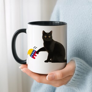 Può includere: Tazza in ceramica bianca con manico e bordo neri. La tazza presenta un'illustrazione di un gatto nero con occhi gialli, che interagisce con un disegno giallo, blu e rosso. La tazza è tenuta da una persona.