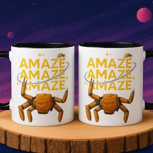 Peut inclure: Deux mugs blancs avec des poignées et des bords noirs, chacun avec une illustration de crabe et le mot "AMAZE" répété trois fois en jaune. Les mugs sont placés sur un fond sur le thème de l'espace.
