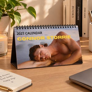 May include: A 2027 desk calendar featuring a photograph of a person lying on a beach. The text 2027 CALENDAR and CONNOR STORRIE is printed in black and yellow on the top portion of the spiral-bound calendar, which is displayed on a wooden surface.