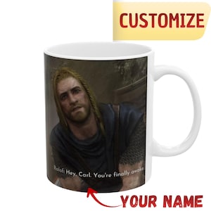May include: White ceramic mug featuring a detailed image of a man with long blonde hair and a beard. The mug has the text "Ralof: Hey, Carl. You're finally awake." and the word "CUSTOMIZE" in a yellow banner.
