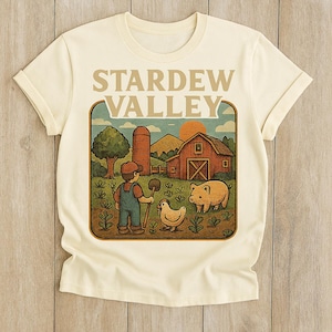 May include: Cream-colored t-shirt featuring a graphic from the video game Stardew Valley. The design includes a farm scene with a barn, silo, farmer, pig, and chicken. The words "STARDEW VALLEY" are printed at the top.