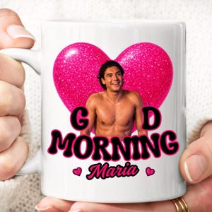 Può includere: Una tazza in ceramica bianca con un grande cuore rosa glitterato. Al centro del cuore c'è la fotografia di un uomo a torso nudo che sorride. Sotto il cuore, il testo nero recita "GOOD MORNING" con il nome "Maria" e due piccoli cuori rosa.