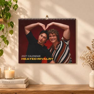 May include: A 2027 wall calendar titled HEATED RIVALRY featuring a photograph of two individuals forming a heart shape with their arms. One person wears a black t-shirt, while the other wears a brown, white, and black striped shirt.