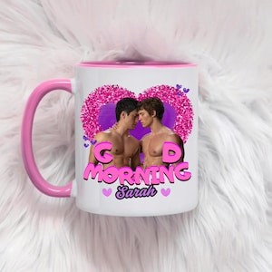 Può includere: Una tazza in ceramica bianca con manico e bordo rosa, caratterizzata da un'immagine di due uomini a torso nudo l'uno di fronte all'altro all'interno di un grande cuore rosa glitterato. Il testo "GOOD MORNING" appare in lettere rosa sotto il cuore, con il nome "Sarah".