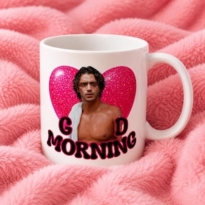 May include: A white ceramic mug featuring a graphic of a person with dark curly hair and a white towel draped over their shoulder, centered in front of a large pink glittery heart. The text "G D MORNING" is printed in black with pink outlines.