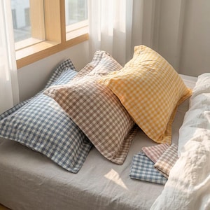 May include: Three square pillows in blue, brown, and yellow gingham patterns are arranged on a bed. Two smaller matching gingham patterned pillowcases are also visible. The pillows are near a window.