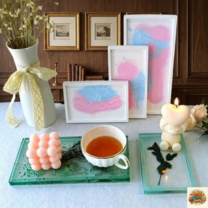 May include: A still-life arrangement featuring a white vase with yellow ribbon, framed art, and various decorative items. Includes a heart-shaped candle, a teacup, and several resin trays with abstract designs in pink and blue.