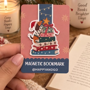 Könnte beinhalten: Ein rechteckiges magnetisches Lesezeichen auf einer rosa Trägerkarte mit dem Text MAGNETIC BOOKMARK und @HAPPYANDGO. Das Lesezeichen zeigt einen Dackel mit Weihnachtsmütze und Pullover, der einen gelben Stern auf einen Bücherstapel setzt.