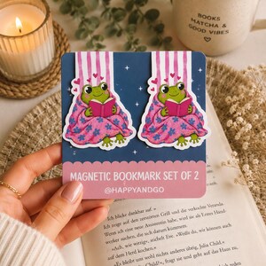 Könnte beinhalten: Ein Set aus zwei magnetischen Lesezeichen mit einem grünen Frosch, der ein rosa Buch liest und in eine rosa Blumendecke eingewickelt ist. Die Lesezeichen sind auf einer dunkelblauen Karte mit dem Text MAGNETIC BOOKMARK SET OF 2 und @HAPPYANDGO befestigt.