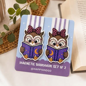 Könnte beinhalten: Ein Set aus zwei magnetischen Lesezeichen mit braunen Eulen, die eine rosa Brille und eine Schleife mit Leopardenmuster tragen und jeweils ein violettes Buch mit Mond- und Sternenmotiven lesen. Die Karte trägt den Text MAGNETIC BOOKMARK SET OF 2 @HAPPYANDGO.