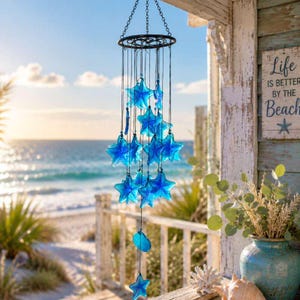 Peut inclure: Un carillon éolien suspendu composé de plusieurs étoiles de mer en verre bleu translucide, fixées à un cadre circulaire en métal noir par de fines cordes noires. L'objet est présenté devant un paysage côtier lumineux avec plage et océan.