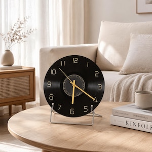 May include: A circular black wall clock crafted from a vinyl record, featuring gold-colored numerals and matching gold-colored hour, minute, and second hands. The clock is supported by a silver-toned metal stand, allowing it to sit upright on a surface.