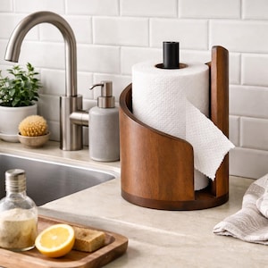 May include: A dark wooden paper towel holder with a curved design. It holds a roll of white paper towels. A black post secures the paper towel roll. The holder is on a kitchen countertop. The holder is made of dark brown wood.