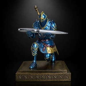 Op de afbeelding: Een metallic blauwe ridderfiguur in volledig harnas knielt op een rechthoekige basis met een decoratieve rand. De ridder heeft gouden en bordeauxrode accenten en houdt een zilveren balpen horizontaal in de handen. Een zwaardgevest is zichtbaar.