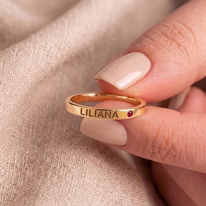 May include: A gold-toned band ring featuring the name LILIANA engraved in black capital letters on the outer surface, accompanied by a small, circular, deep red gemstone set flush into the metal band next to the engraved name.
