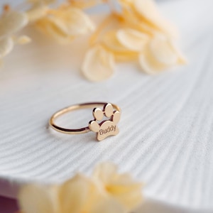 May include: A gold-colored ring featuring a paw print design with the name "Buddy" engraved on it. The ring is set against a white, textured surface, with soft, blurred floral accents in the background.