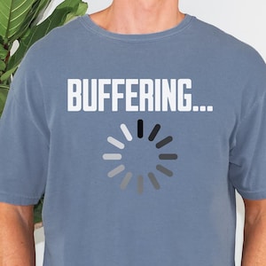 May include: A blue short-sleeved t-shirt featuring the white text "BUFFERING..." above a circular loading icon composed of twelve gray segments arranged in a radial pattern. The shirt has a crew neckline and a relaxed fit, paired with gray denim jeans.