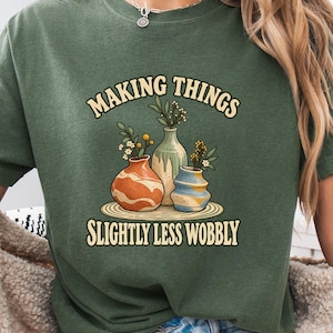 May include: A moss green short-sleeve t-shirt featuring a graphic of three ceramic vases on a mat with the text "MAKING THINGS SLIGHTLY LESS WOBBLY" in a curved font. The vases are orange, cream, and blue with small floral sprigs inside them.