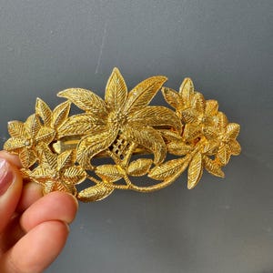 Könnte beinhalten: Eine goldfarbene Haarspange aus Metall mit einem detaillierten Blumendesign aus mehreren übereinanderliegenden Blütenblättern und Blättern. Die Metalloberfläche hat ein strukturiertes, geätztes Finish, das das Licht einfängt und ein helles, reflektierendes Erscheinungsbild erzeugt.