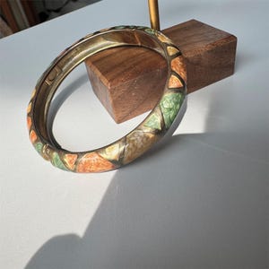 May include: A hinged bangle bracelet featuring a metallic frame with inlaid segments of marbled enamel in shades of green, orange, and cream. The bracelet has a smooth, rounded profile and a visible hinge mechanism on the side for opening and closing.