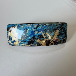 May include: A rectangular hair barrette featuring a glossy finish with a marbled pattern in shades of deep blue, black, and bright gold leaf accents. The accessory has a smooth, curved surface and a polished appearance throughout the design.