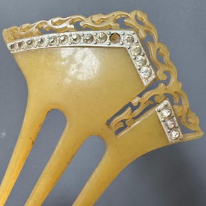 May include: A vintage hair comb with a light amber color. The comb features a decorative silver-toned border adorned with clear rhinestones. The design includes ornate, swirling patterns, and the comb has three long, slender teeth.