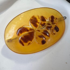 Op de afbeelding: Ovale, amberkleurige broche met een klaverbladontwerp. De klaver heeft een schildpadpatroon met donkerbruine vlekken. De broche heeft een metalen speld op de achterkant.