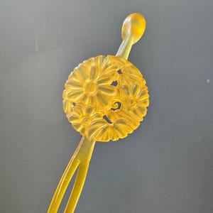 Può includere: Un pettine giallo per capelli con un design floreale. Il pettine presenta una disposizione floreale circolare scolpita con uno stelo e una parte superiore arrotondata. Il pettine è realizzato in un materiale giallo traslucido.