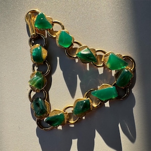 May include: A gold-tone chain bracelet featuring a series of interconnected circular links, each adorned with a polished, irregular-shaped green stone set within a textured, diamond-shaped gold metal frame, secured by a cylindrical bar clasp.