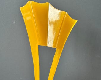 Puede incluir: Un accesorio para el cabello de color amarillo brillante con una parte superior en forma de abanico con tres crestas verticales distintas y dos púas largas y delgadas que se extienden hacia abajo. El objeto tiene un acabado liso y brillante y un aspecto translúcido.