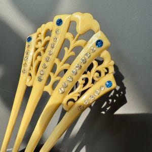 May include: Ivory-colored hair sticks with intricate openwork designs. Each stick is adorned with sparkling clear rhinestones and a single blue gemstone. The hair accessories are arranged in a fan-like pattern, showcasing their ornate details.