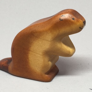 May include: A wooden beaver figurine in shades of brown. The beaver is in a seated position, with its paws clasped together. The tail is flat and paddle-shaped. The figurine has a smooth finish and simple, painted facial details.