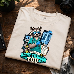 May include: A beige t-shirt featuring a graphic of a raccoon dressed as a radiologist wearing blue sunglasses, a teal scrub top, and blue gloves. The raccoon holds an X-ray film and a clipboard with the text RADIOLOGY. The text reads I CAN SEE RIGHT THROUGH YOU.