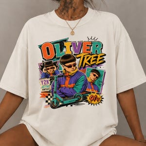 May include: An off-white short-sleeve t-shirt featuring a graphic print of Oliver Tree with a teal scooter. The design includes the text "OLIVER TREE", "Y2K RETRO VINTAGE", and "90s" in colorful, bold lettering with various geometric shapes and patterns.