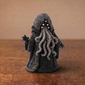 Op de afbeelding: Een grijze vilten Cthulhu-beeldje met een capuchon en tentakels. Het beeldje heeft zes witte stippen op het hoofd en staat op een houten oppervlak. De armen van het beeldje zijn uitgestrekt.