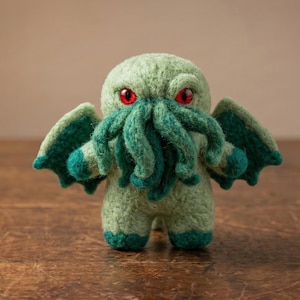Könnte beinhalten: Handgefertigtes, grünes Cthulhu-Plüschtier mit roten Augen und türkisfarbenen Tentakeln und Flügeln. Das Plüschtier besteht aus einem weichen, strukturierten Material und steht auf einer Holzoberfläche. Die Kreatur hat ein skurriles, cartoonartiges Aussehen.