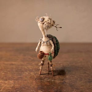 May include: A handcrafted creature figurine with a pale, textured body, a dark green shell, and a wide, toothy grin. It features a small brown pouch and a green sack attached to a red waist cord, with thin, stick-like legs and wispy hair.