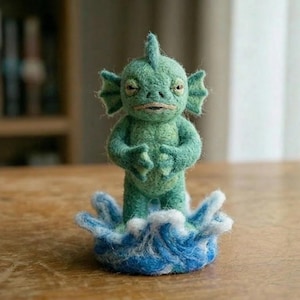 May include: A needle-felted creature figurine in shades of green, featuring a dorsal fin, side fins, and a wide mouth. The creature stands upright on a base crafted to resemble a circular splash of blue and white water, with a textured surface.