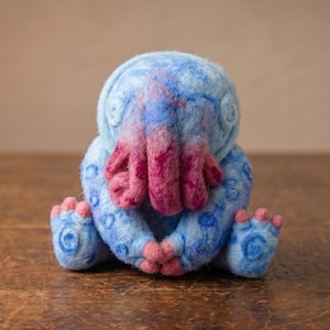 May include: A needle-felted creature figurine featuring a light blue body with darker blue swirl patterns and a cluster of pink and magenta tentacles covering its face. The creature has small pink claws on its hands and feet, with a rounded head.
