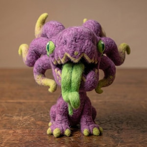 May include: A handmade felt creature in shades of purple and green. The monster has multiple tentacles, green eyes, and a long green tongue. The creature is sitting on a wooden surface.