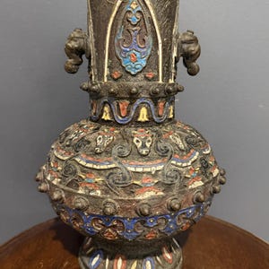 May include: A dark bronze-toned metal vase featuring intricate relief carvings of animal heads and geometric patterns. The surface is accented with enamel in shades of blue, red, yellow, and white. It has two side handles and a wide, rounded body.