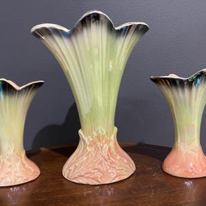 Puede incluir: Tres jarrones de cerámica con diseño floral. Los jarrones tienen una parte superior acampanada y con volantes con un borde iridiscente oscuro. Los cuerpos son de color verde pálido con una base rosa. El jarrón más grande mide unos 25 cm de alto.