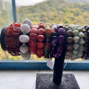 May include: A selection of beaded bracelets in a range of colours, including red, white, purple, and green. The bracelets are made from various materials and are displayed on a black stand. A window with a mountain view is in the background.