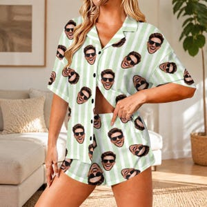 May include: A two-piece pajama set consisting of a short-sleeved button-down shirt and matching shorts. The fabric features a light green and white vertical striped pattern overlaid with a repeating print of a smiling face wearing sunglasses.