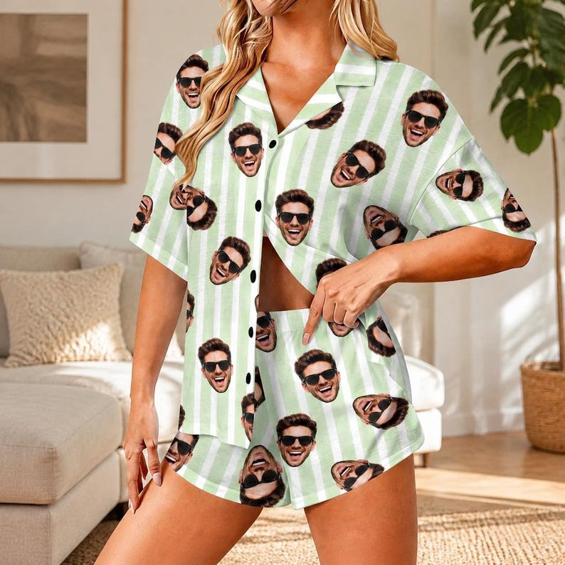 May include: A two-piece pajama set consisting of a short-sleeved button-down shirt and matching shorts. The fabric features a light green and white vertical striped pattern overlaid with a repeating print of a smiling face wearing sunglasses.