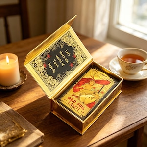 Puede incluir: Un mazo de cartas de tarot con lámina dorada en una caja dorada abierta. La tapa de la caja tiene el texto "Let's share the world of Tarot happily" en negro y dorado. Las cartas son visibles dentro de la caja, con detalles e ilustraciones en lámina dorada. Una vela encendida y una taza de té están en el fondo.