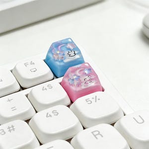 May include: Close-up of a white keyboard with two custom keycaps. One keycap is blue with a smiling cloud and stars, the other is pink with a smiling moon and stars. The keycaps are translucent and have a glittery effect.