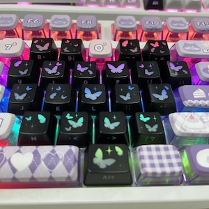May include: Close-up of a mechanical keyboard with custom keycaps. The keycaps are black with butterfly designs in various colors, including blue, purple, and green. Some keys have cat and cake designs. The keyboard has a white frame.