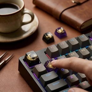 Può includere: Primo piano di una tastiera meccanica con tasti personalizzati. I tasti presentano disegni oro e neri con una base viola traslucida. Un dito preme un tasto. Una tazza di caffè e un taccuino sono sullo sfondo.