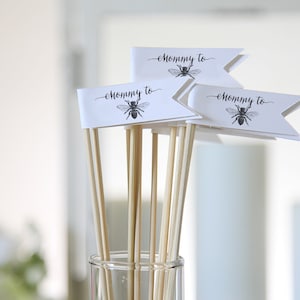 Może przedstawiać: Trzy białe flagi z czarnymi ilustracjami pszczół i tekstem "Mommy to" na każdej fladze. Flagi są przymocowane do drewnianych patyczków i umieszczone w przezroczystym szklanym wazonie.